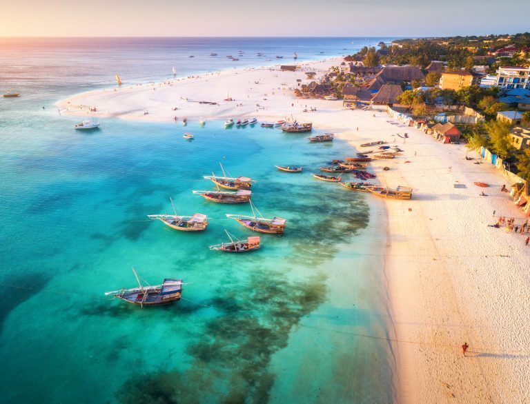 Aerial view of the fishing boats on tropical sea coast with sandy beach at sunset. Summer holiday on Indian Ocean, Zanzibar, Africa. Landscape with boat, buildings, transparent blue water