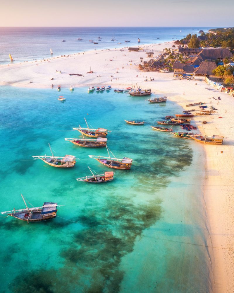 Aerial view of the fishing boats on tropical sea coast with sandy beach at sunset. Summer holiday on Indian Ocean, Zanzibar, Africa. Landscape with boat, buildings, transparent blue water. Top view